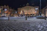 a city with cobblestones and buildings at night, lights are on, but a fountain is in the middle