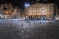 a city with cobblestones and buildings at night, lights are on, but a fountain is in the middle