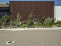 a street lined with plants and bushes in front of a brown building under a blue sky