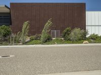 a street lined with plants and bushes in front of a brown building under a blue sky