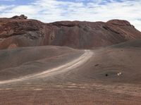 an empty dirt road cuts through the desert near a large hill with trees and rocky cliffs