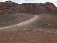 an empty dirt road cuts through the desert near a large hill with trees and rocky cliffs