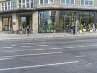 people standing outside a bus stop by the road with windows filled in pots, and bicycles parked nearby