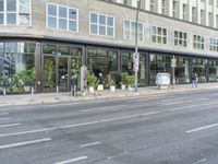 people standing outside a bus stop by the road with windows filled in pots, and bicycles parked nearby