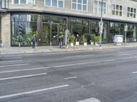 people standing outside a bus stop by the road with windows filled in pots, and bicycles parked nearby