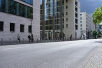 people walking on the side of a road by tall buildings near some street lights and trees