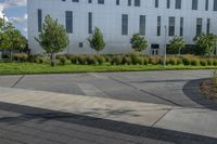 the man is riding his skateboard on the sidewalk near the building with trees, plants and bushes