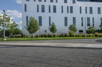 the man is riding his skateboard on the sidewalk near the building with trees, plants and bushes