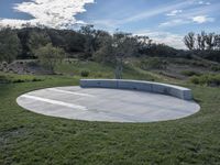 a concrete seating area sitting in a grassy field next to a hillside of trees on a sunny day