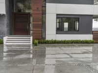 a white fence on the sidewalk in front of a house in the rain and some green bushes