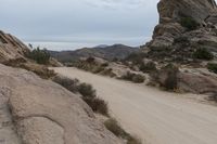 a dirt road runs between rocky cliffs and scrub brush with shrubbery near it while the sky is white