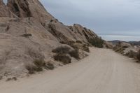 a man riding a motorcycle down a dirt road in the desert near some rocks and bushes