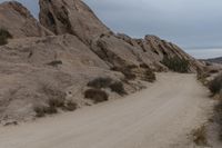 a man riding a motorcycle down a dirt road in the desert near some rocks and bushes