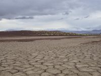 the barren field has dry mud and trees in it under cloudy skies above mountains with plants on them