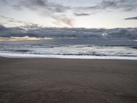 a sandy beach and some ocean waves at dusk, with clouds above the water,