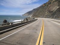 an empty highway next to the ocean with some rocks on it with blue sky in background