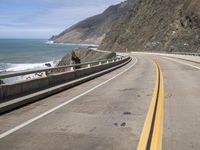 an empty highway next to the ocean with some rocks on it with blue sky in background