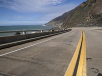 an empty highway next to the ocean with some rocks on it with blue sky in background