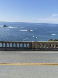 an older couple rides their bicycle on the bridge overlooking the ocean and coastline below a blue sky