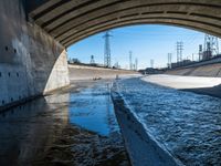 water flowing under an overpass with power lines in the background and overhead wires and a bridge