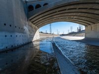 water flowing under an overpass with power lines in the background and overhead wires and a bridge