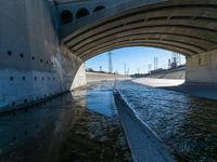 water flowing under an overpass with power lines in the background and overhead wires and a bridge