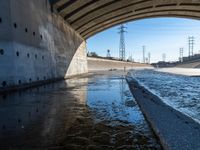 water flowing under an overpass with power lines in the background and overhead wires and a bridge