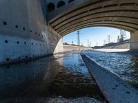 water flowing under an overpass with power lines in the background and overhead wires and a bridge