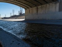 water flowing under an overpass with power lines in the background and overhead wires and a bridge