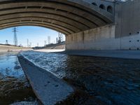 water flowing under an overpass with power lines in the background and overhead wires and a bridge