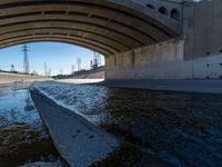 water flowing under an overpass with power lines in the background and overhead wires and a bridge