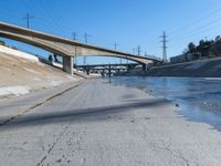 a narrow road with puddles on the ground, underneath an overpass and many electrical wires