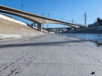 a narrow road with puddles on the ground, underneath an overpass and many electrical wires