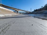 a narrow road with puddles on the ground, underneath an overpass and many electrical wires