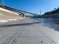 a narrow road with puddles on the ground, underneath an overpass and many electrical wires