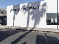 a white building with black letters on the side and shadow on the pavement with palm trees