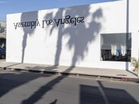 a white building with black letters on the side and shadow on the pavement with palm trees