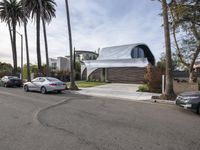 a white house has a sloping roof on the street by some palm trees and parked cars