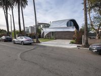 a white house has a sloping roof on the street by some palm trees and parked cars