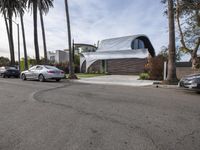 a white house has a sloping roof on the street by some palm trees and parked cars