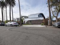 a white house has a sloping roof on the street by some palm trees and parked cars