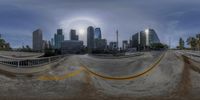 this is a fish eye lens image of a half pipe skatepark at the base of a city in the background