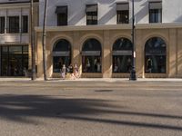 people walk along a street past a building and shops with arched windows on both sides