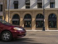 a red car passes by the building with arches along the street as a traffic light