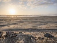 a sunset over the beach as people walk on sand and rocks in the water are visible