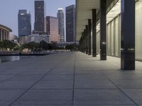 the walkway at the park leads to a lake and buildings in the background as evening falls