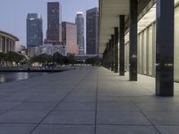 the walkway at the park leads to a lake and buildings in the background as evening falls