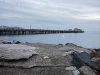 rocks on the beach next to a body of water with a pier in the background