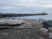 rocks on the beach next to a body of water with a pier in the background