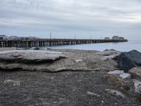 rocks on the beach next to a body of water with a pier in the background